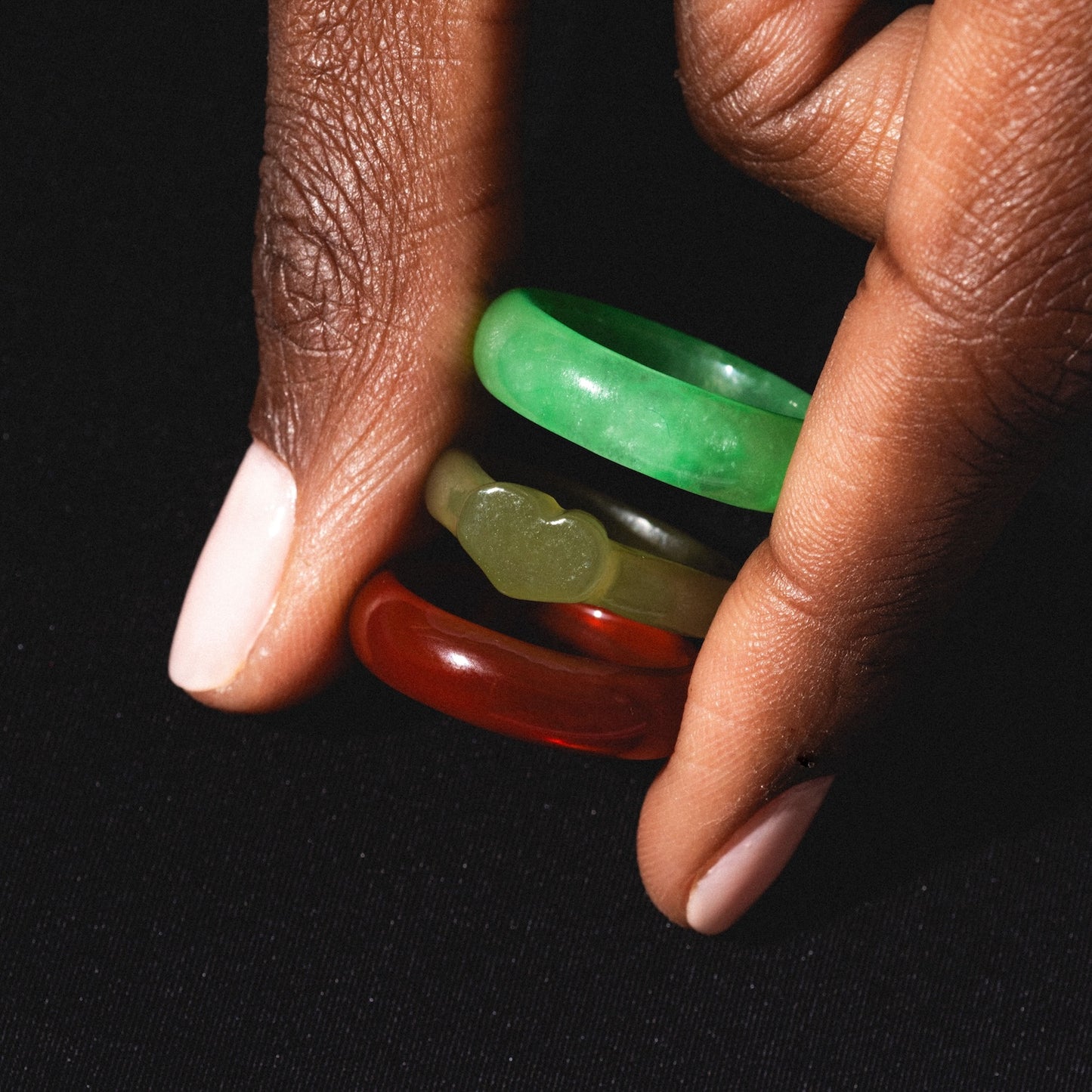 A hand with light pink polish holds three Heart Jade Rings—green, red, and a heart-shaped one—each with a 3.5 mm band width, against a black background.