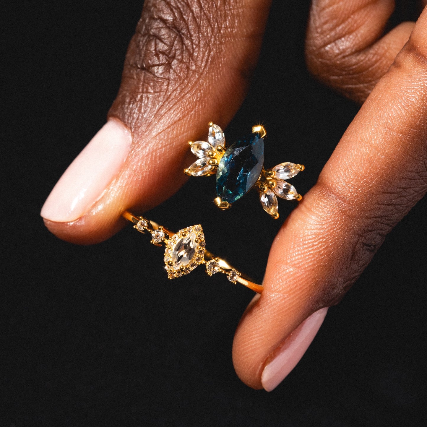 A hand with pale pink polished nails holds two gold rings—one is the White Topaz Harlow Ring in 18k gold vermeil, featuring a sparkling marquise white topaz and clear accents—set against a black background.