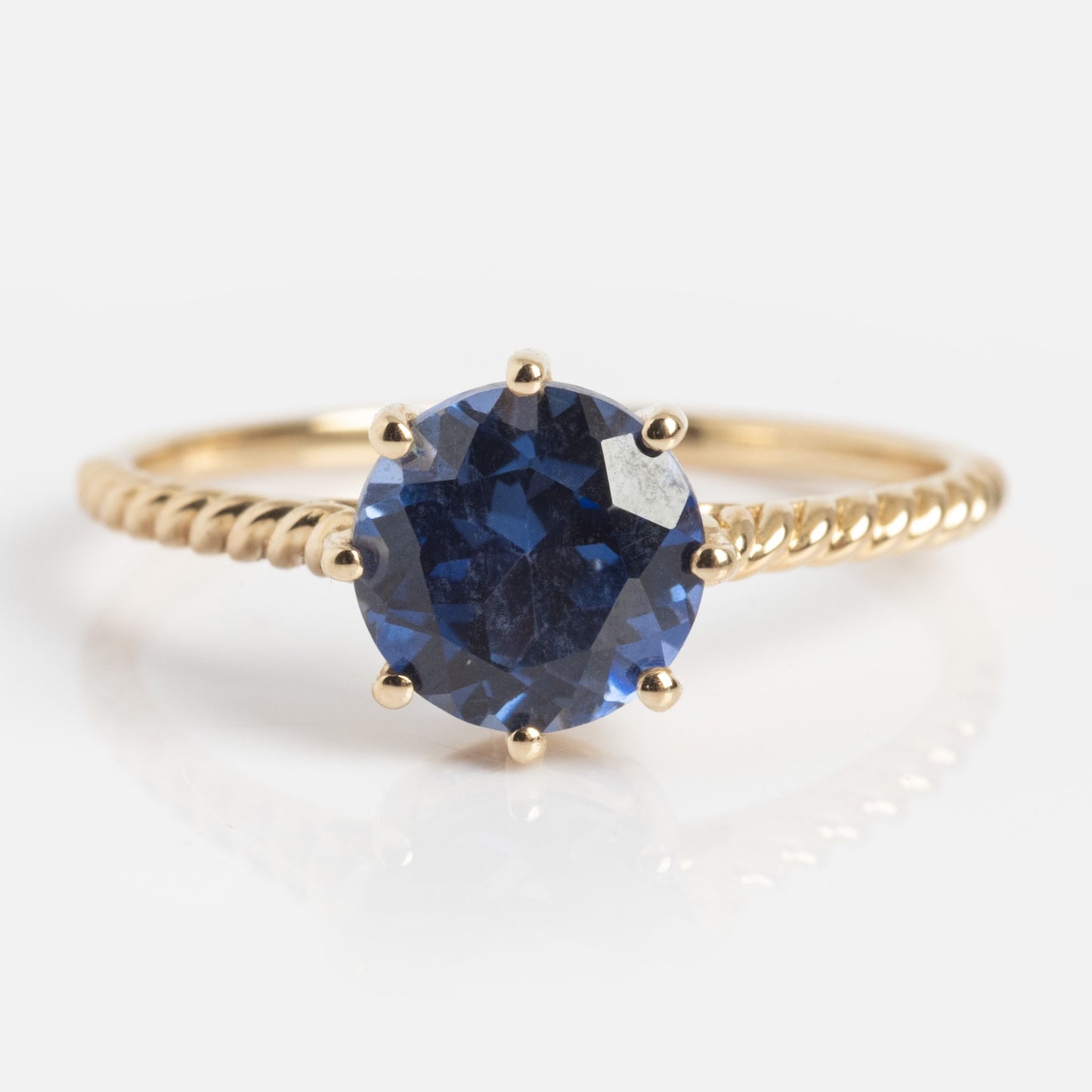 The Solid Gold 2025 December Capsule Classic Tanzanite Statement Ring showcases a round, faceted deep blue tanzanite in a six-prong setting atop a twisted gold band, displayed on a white background.