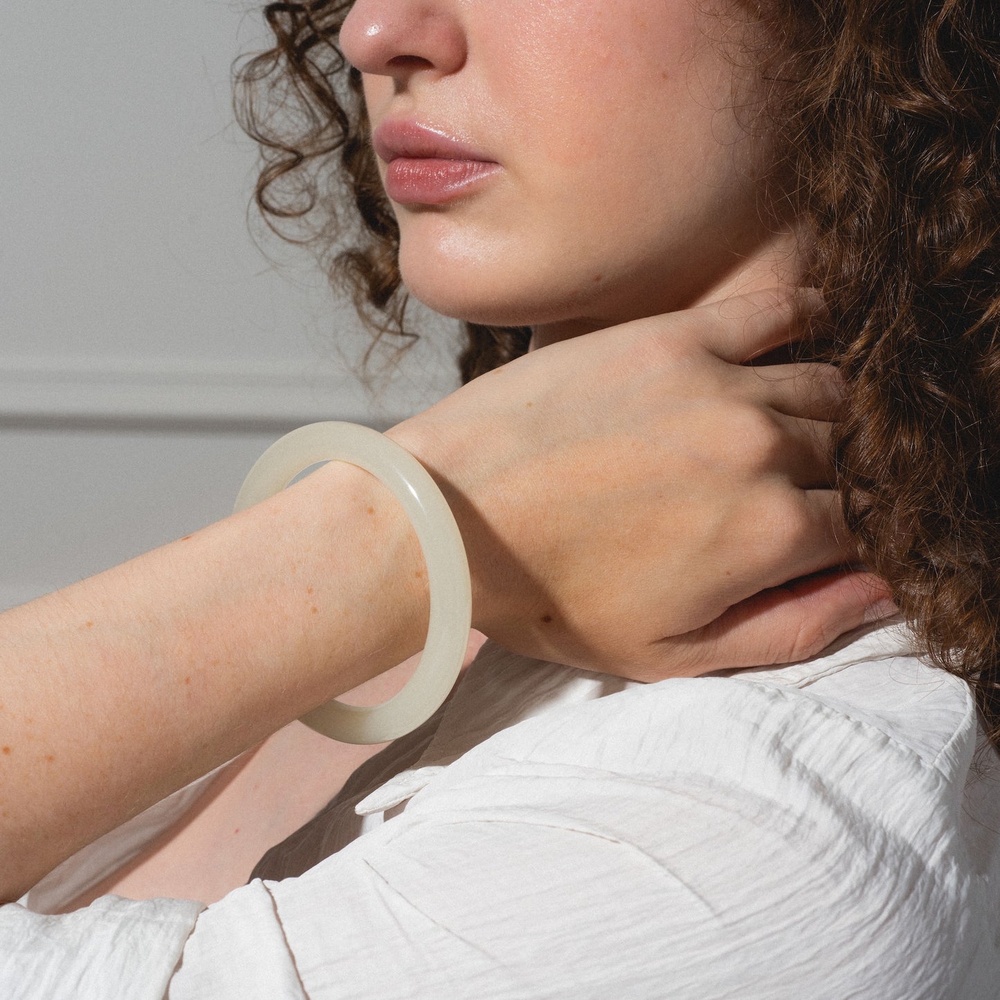 A woman in a white shirt wears the 1994 Off White Thin Jade Bangle on her wrist, touching her neck. She has curly brown hair, natural makeup, and is photographed from the shoulders up against a plain background.