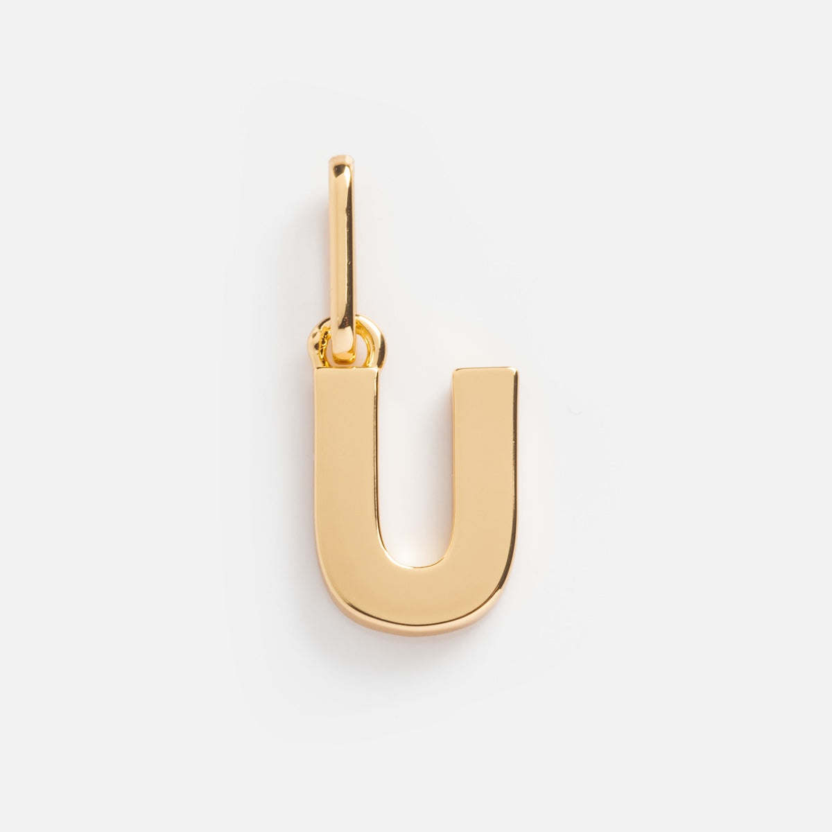 The Be You Initial Charm, shaped like the letter U in gold, hangs from a small loop and highlights elegant personalized jewelry on a plain white background.