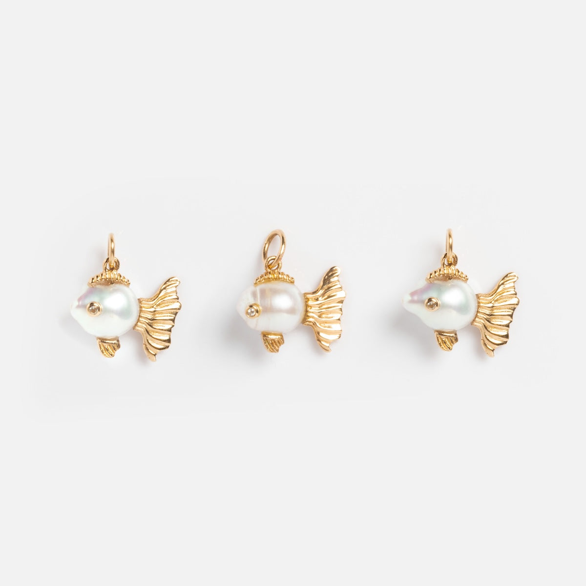 Three Solid Gold Nature Charms are arranged in a row on a plain white background. Each features a fish shape with gold fins, a pearl body, and a small loop at the top for attaching to jewelry.
