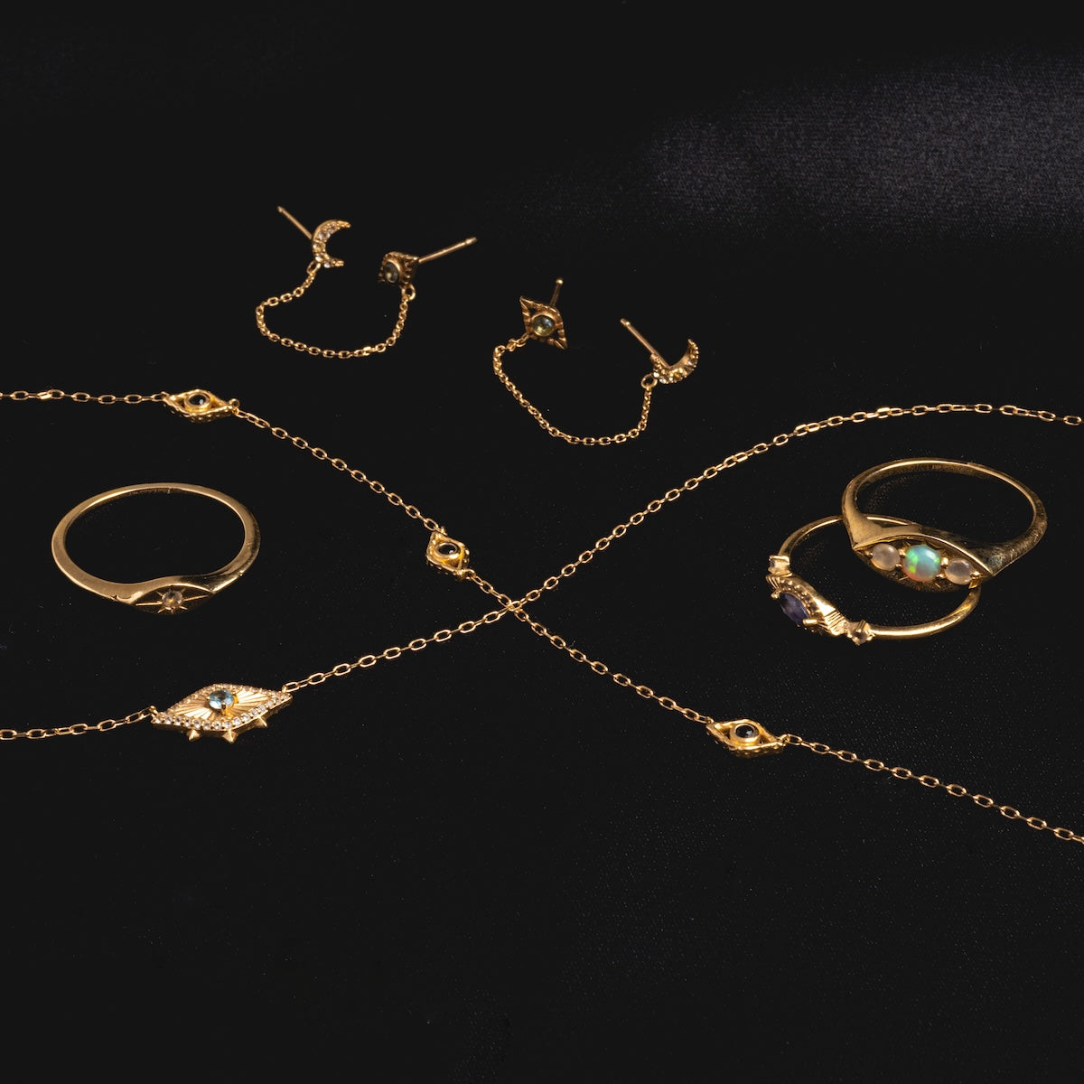 Gold rings, chain bracelets, and hoop earrings with gemstone and star designs are arranged on black satin fabric. Intricate details like crescent moons and the Solid Gold Opal Evil Eye Signet Ring create a captivating display.