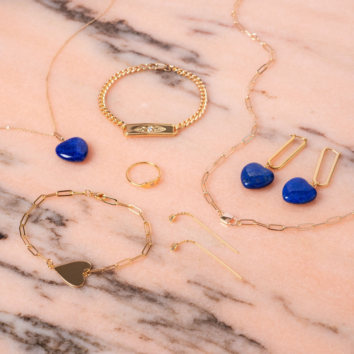 The Divine Feminine Bracelet, along with gold-plated necklaces, rings, earrings, and blue heart-shaped stone pendants, is displayed on a pink marble surface.