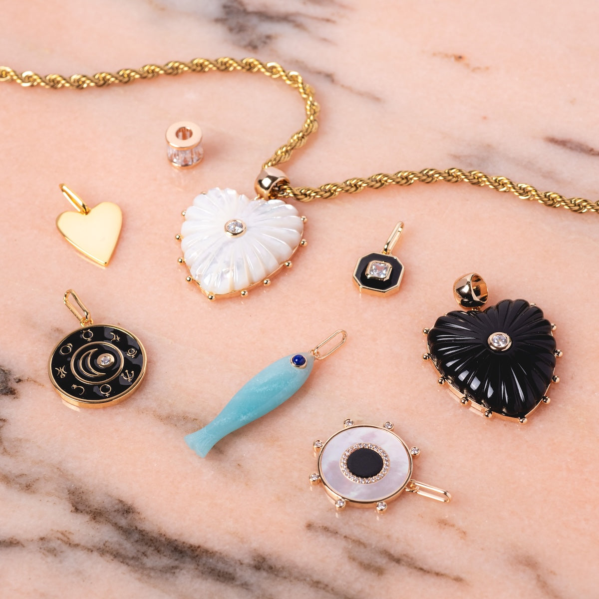 Charms by Thatch features gold plated and enamel charms—heart shapes, a blue fish, a round camera, and a mother of pearl charm—arranged on pink marble for unique personalized necklace options.