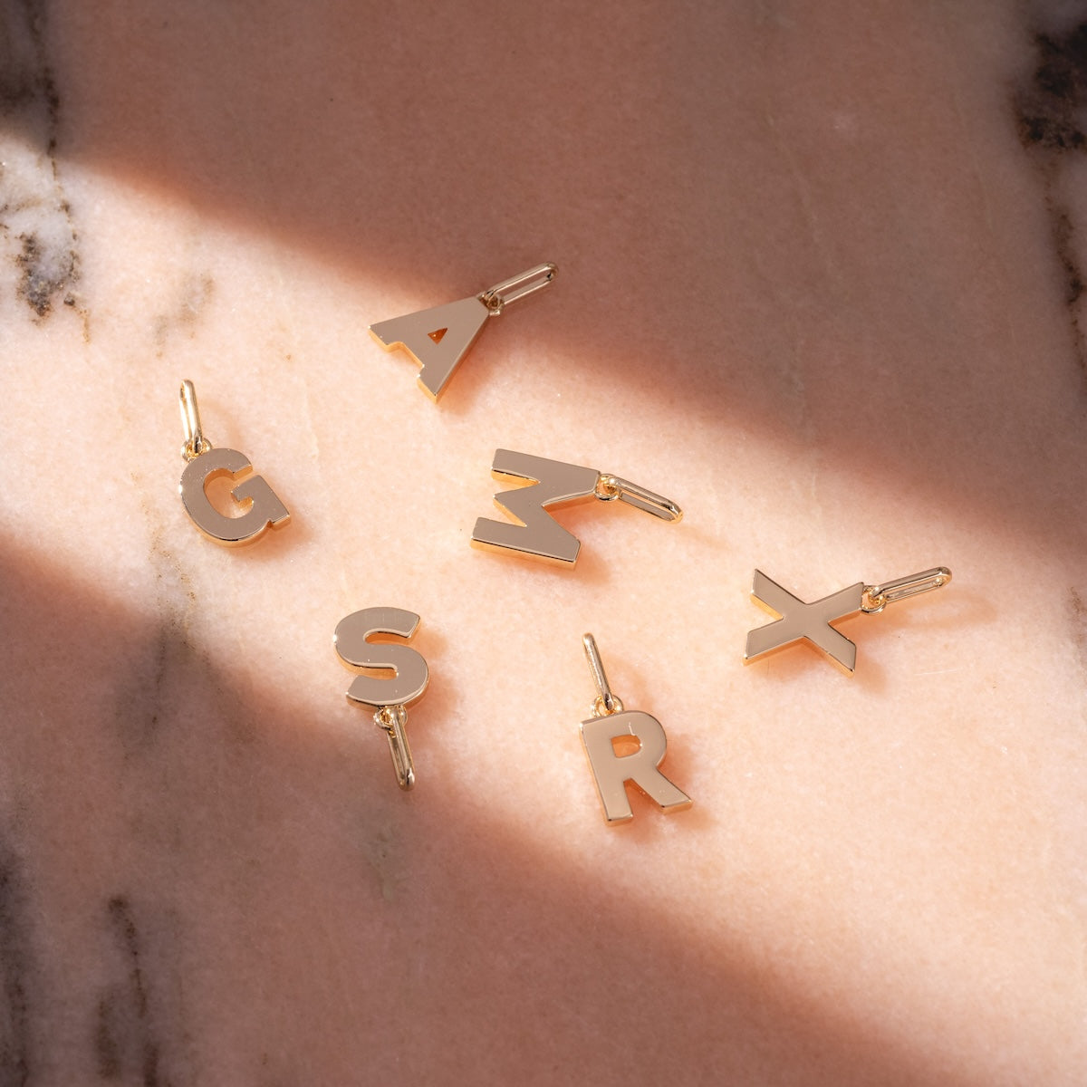 Be You Initial Charms in gold, featuring the letters G, S, A, R, M, and X, are arranged on pink marble. Sunlight casts elegant shadows across these personalized jewelry pieces for a striking display.