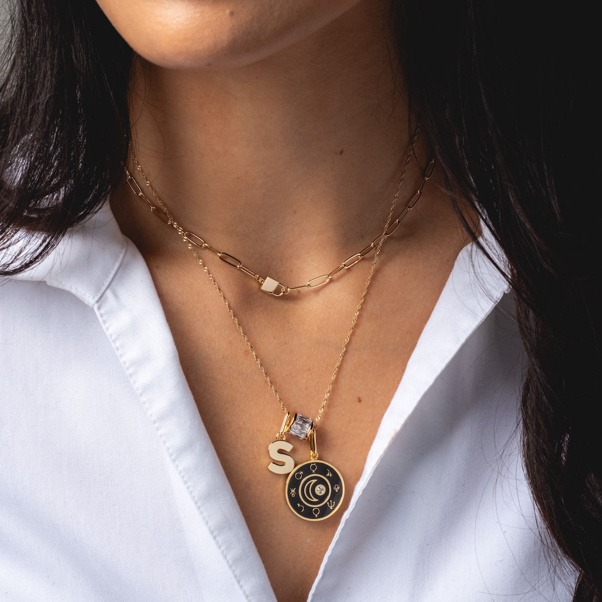 A woman wears a white collared shirt and two gold Charms by Thatch necklaces—one with a large S pendant, plus personalized charms: a clear gemstone and a round black charm with gold crescent moon and stars.