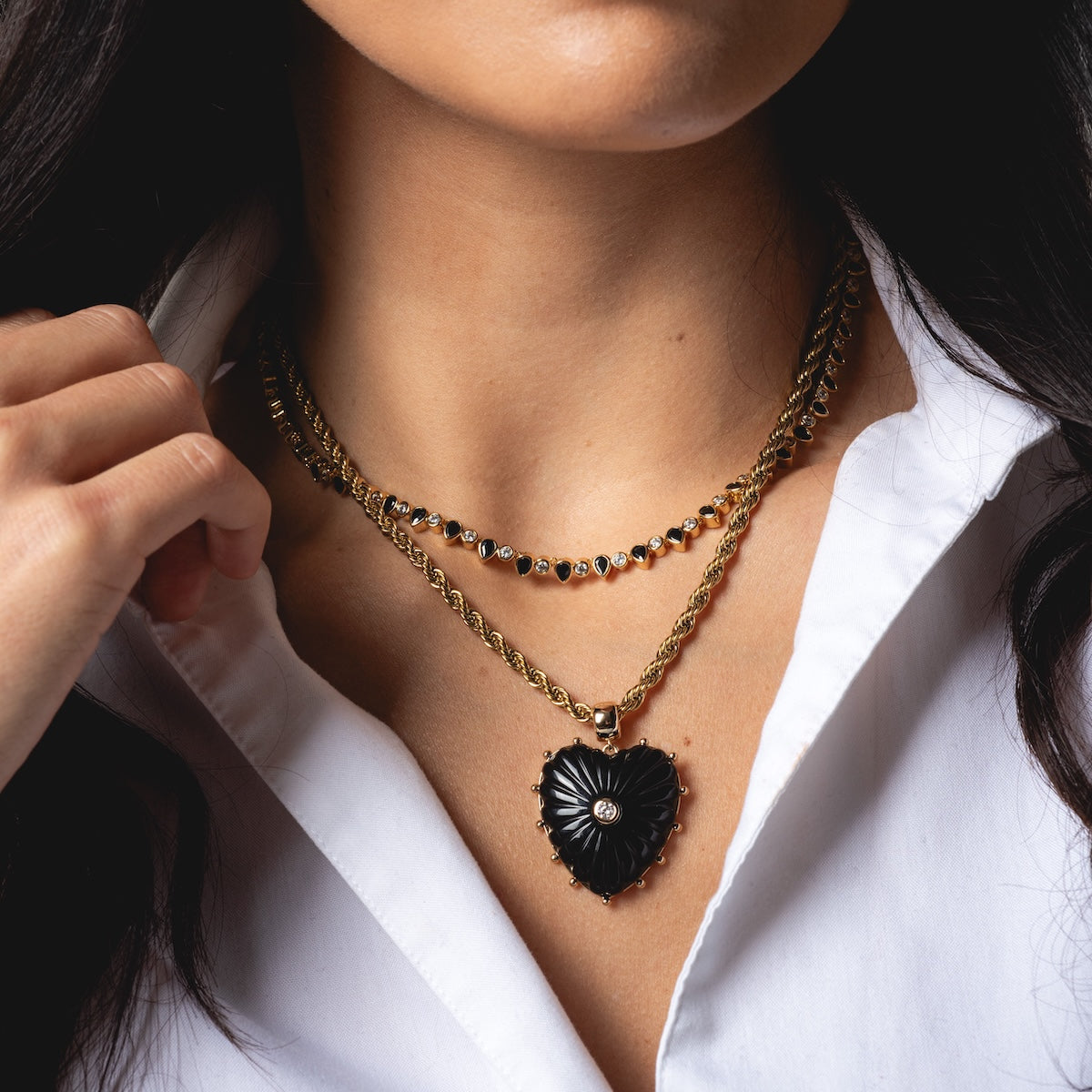 A woman in a white blouse displays Charms by Thatch layered gold necklaces—one with small black stones, another with a large black heart pendant—and touches a necklace featuring elegant gold plated charms by her neck.