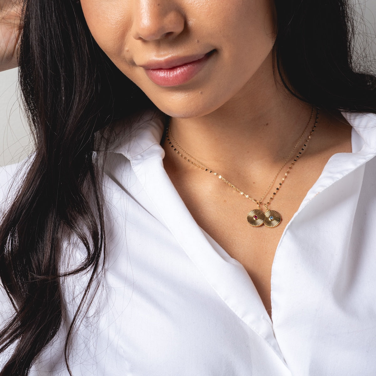 A woman smiles softly, wearing a white button-up shirt and two 14k gold plated Mantra Empowerment Coin Charm necklaces. The image highlights her long dark hair and focuses on the empowering jewelry at her neck and chest.