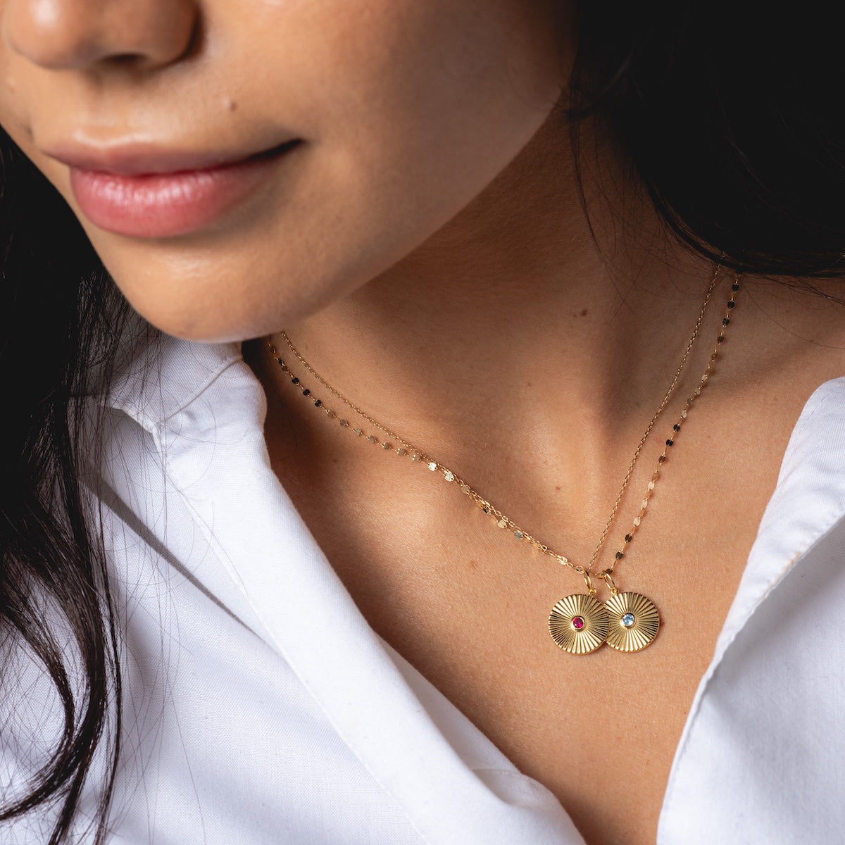 A woman wears a white shirt and two layered 14k gold plated Mantra Empowerment Coin Charm necklaces with round, textured pendants featuring small red and white gemstones. Only her lower face and part of her neck are visible.