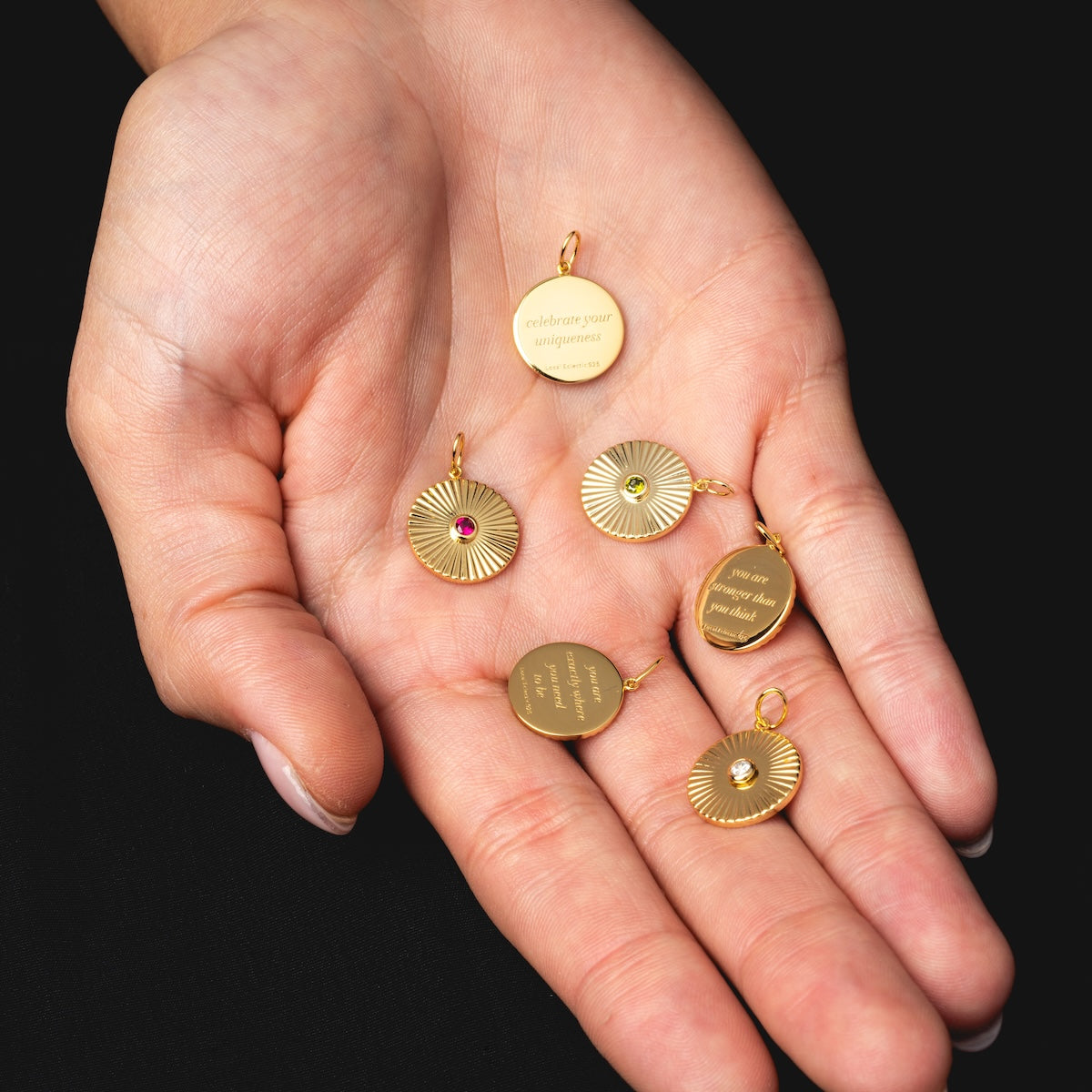 A hand holds six Mantra Empowerment Coin Charms, 14k gold plated pendants engraved with uplifting text and accented with tiny gemstones—ideal mantra jewelry for those seeking inspiration, shown against a black background.