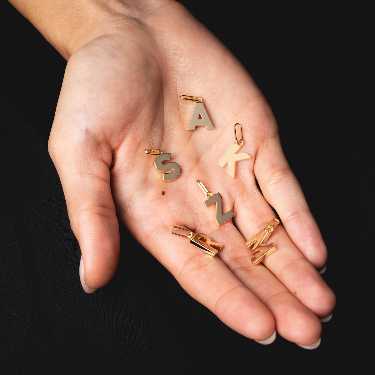 A hand displays six Be You Initial Charms—gold letters S, A, K, S, N, and M—against a black background, ideal for creating personalized gold necklaces or custom jewelry.