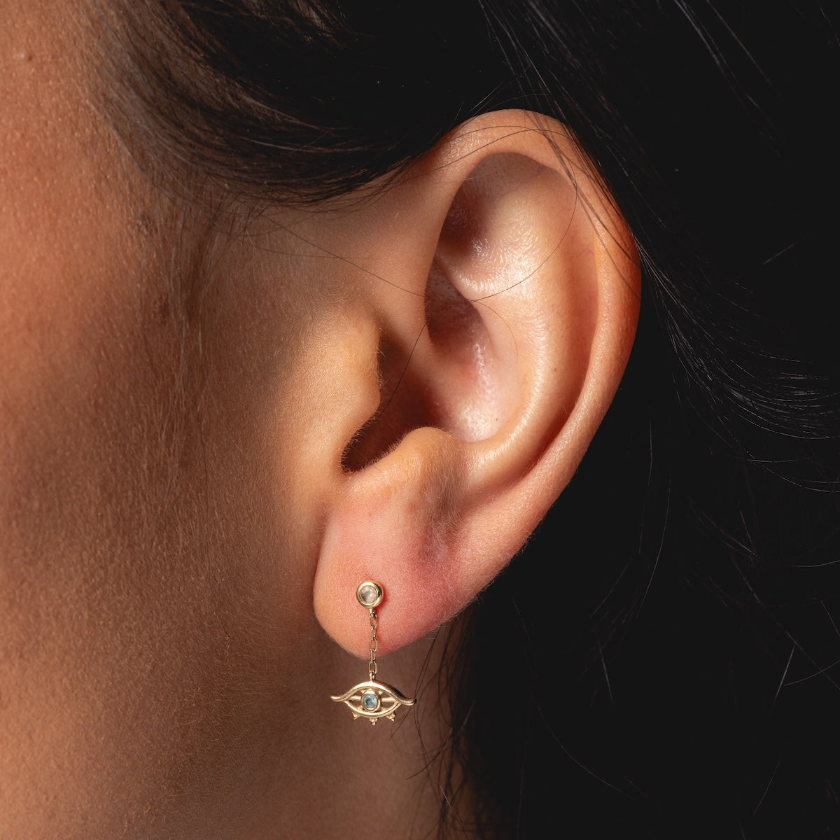 Close-up of a person’s ear wearing Harvest Moon Jewelry’s Solid Gold Evil Eye Drop Studs, featuring a London Blue Topaz at the center and a small gemstone above. The person’s dark hair is pulled back.