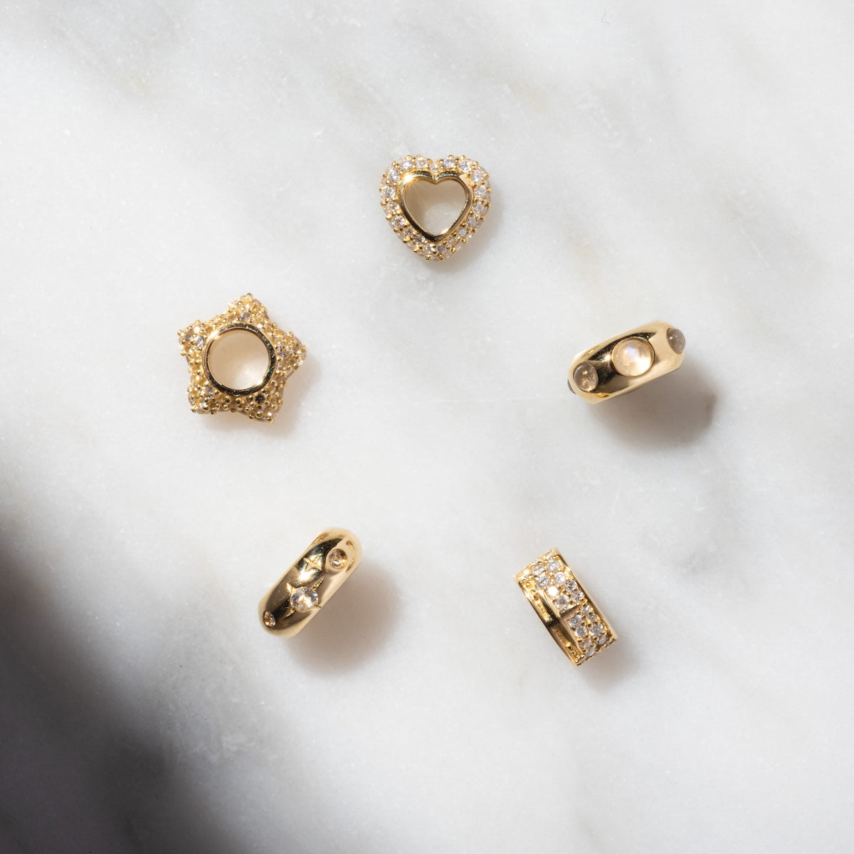 Five Solid Gold Spacer Charms, including heart and star shapes—some with clear crystals—are displayed on white marble with light and shadow, making them ideal gemstone spacer charms for your unique jewelry designs.