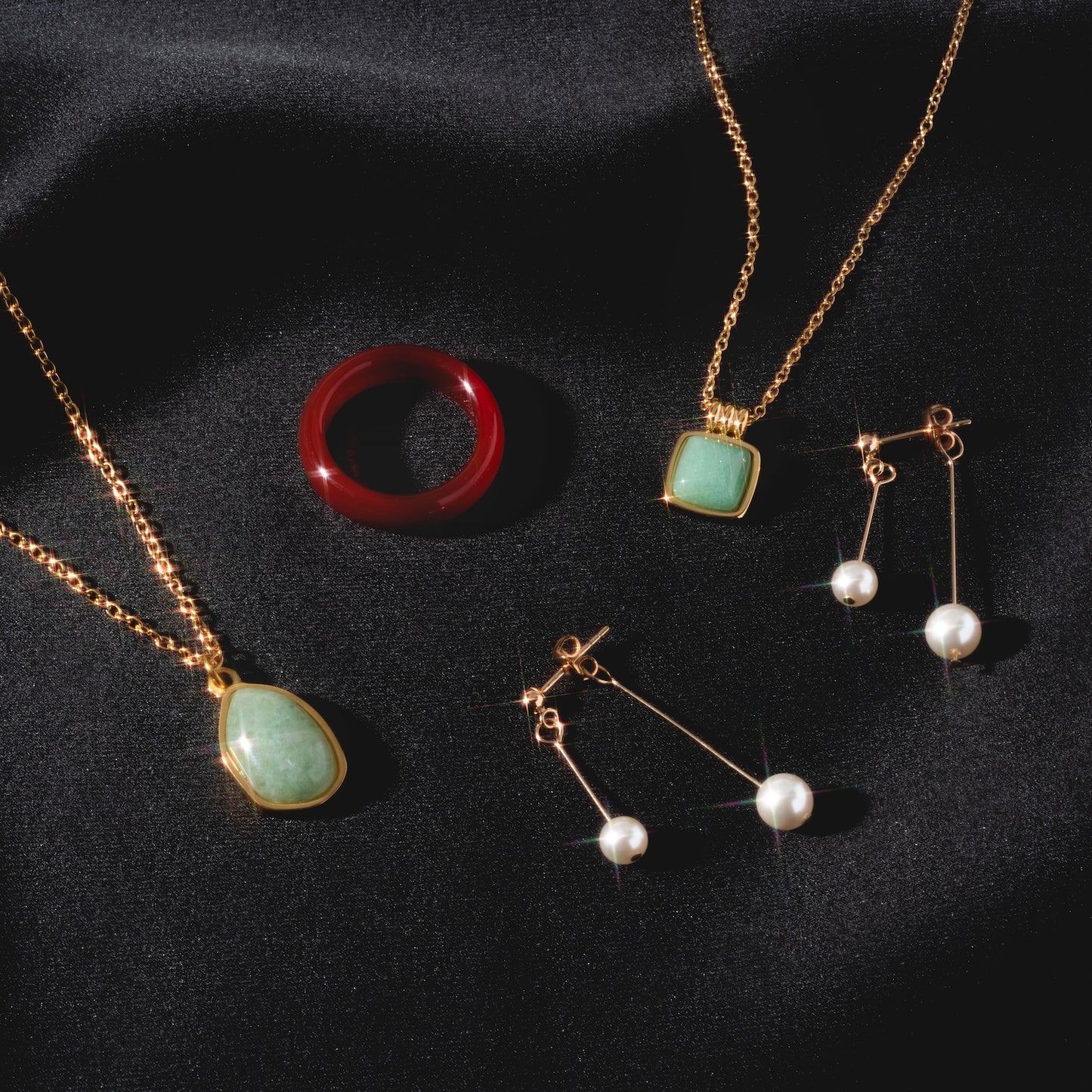 The Puzzle Square Aventurine Stone Necklace, a red ring, and two pairs of pearl drop earrings are displayed on black satin fabric under soft lighting.
