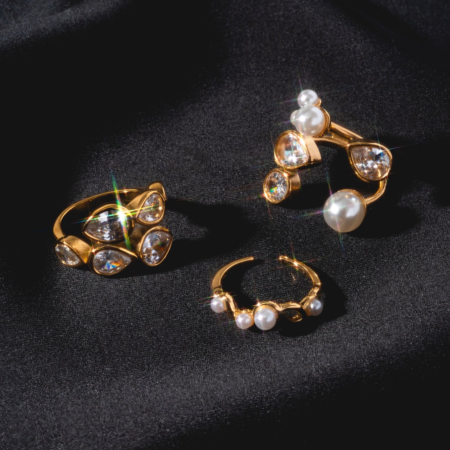 The Pearl and Clear Wrap Ring, plated in 18k yellow gold with sparkling gemstones and pearls, is displayed on black satin fabric—its elegant design and radiant shine make it a stunning statement piece.