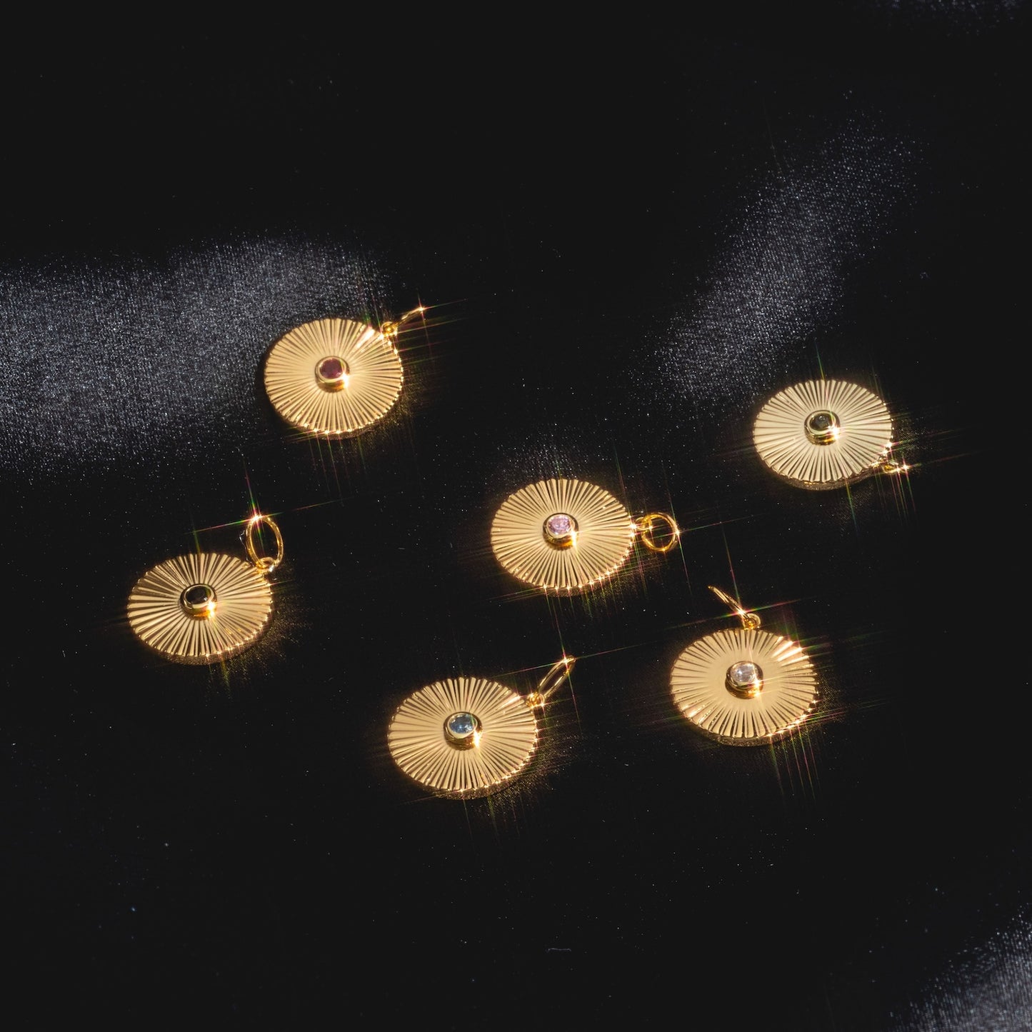 Five round Mantra Empowerment Coin Charms, featuring 14k gold plating with radiating lines and a central gemstone, are displayed on black fabric, reflecting light with star-like sparkle—ideal as mantra jewelry or an empowerment charm.
