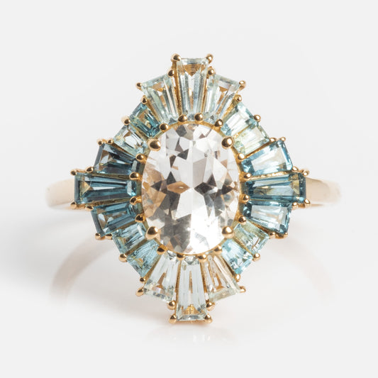 The 14k Bejeweled Blue Statement Ring showcases a large oval clear center stone with a sunburst halo of baguette-cut blue and light blue gems—an eye-catching gold ring for bold style.