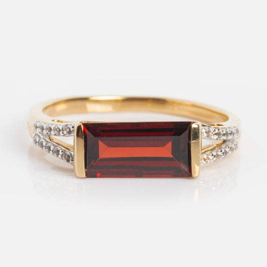 The 14k Gilded Garnet East West Baguette Ring features a rectangular red garnet set horizontally at the center, with rows of clear accent stones on each side of the solid gold band, shown against a white background.