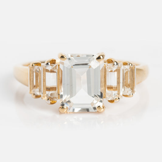 The 14k Graduated White Topaz Engagement Ring features a large emerald-cut centerpiece and three smaller rectangular white topaz stones on each side, all set in solid gold for a classic, elegant look.