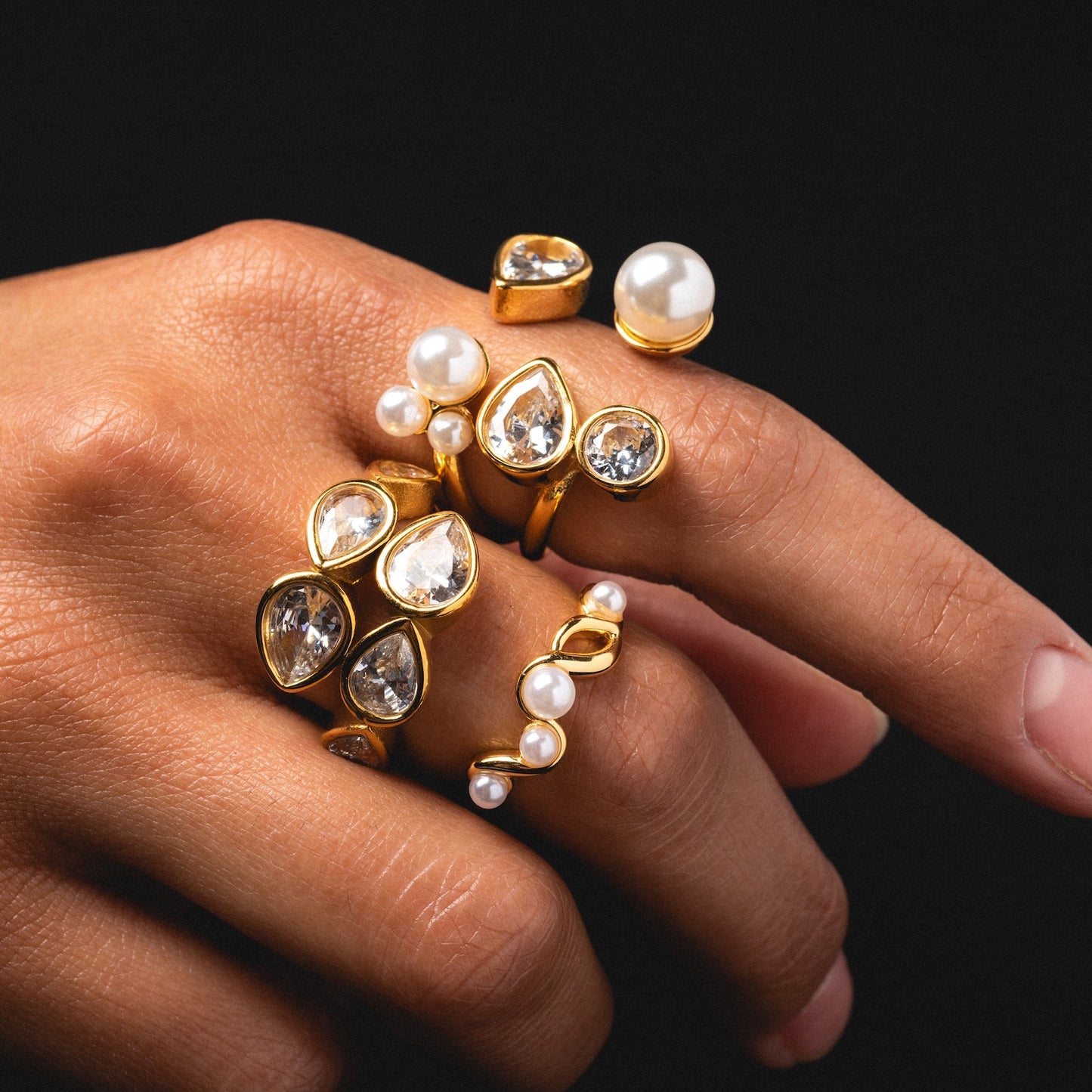 A hand displays the Pearl and Clear Wrap Ring, an 18k yellow gold plated ring with pearl and crystal accents, alongside other modern designs across several fingers against a black background.