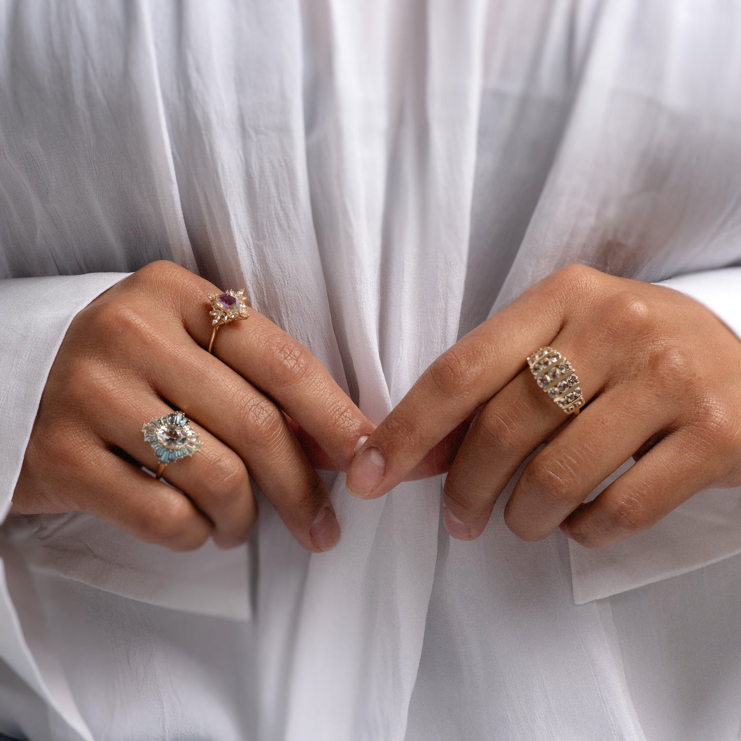 A person in a sheer white shirt wears three ornate rings: a diamond-studded 14k gold ring, the 14k Bejeweled Blue Statement Ring, and a ring featuring a pink stone.