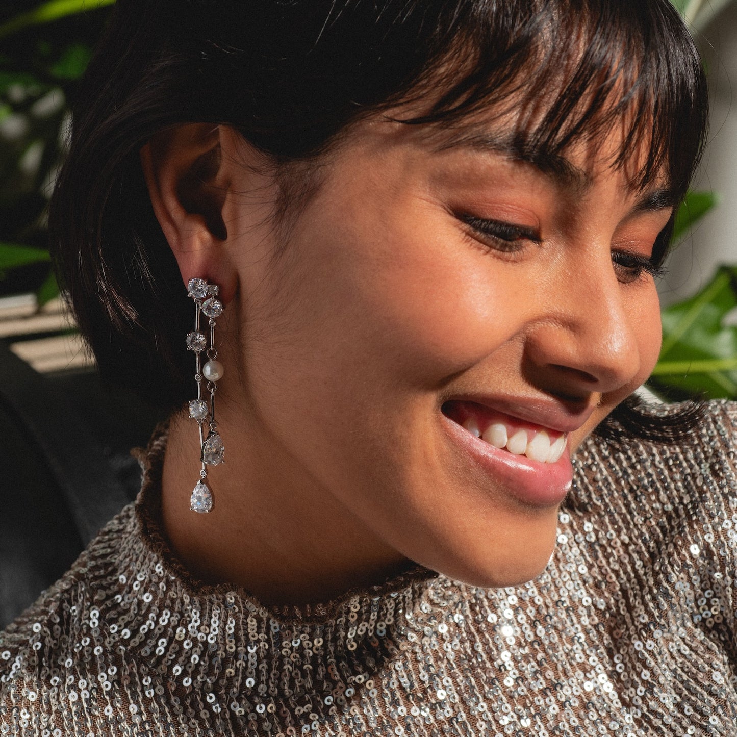 A woman smiles and looks down, wearing a sparkly sequin top and the Jones Diamond Drop Earrings Sample. Sunlight highlights her face and jewelry, with green plants softly blurred in the background.