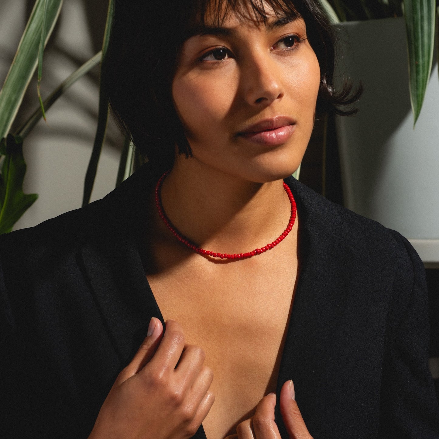A woman with short dark hair in a black blazer and the Rojo Necklace Sample gazes thoughtfully to the side. Green plants are behind her, and warm light highlights her face and necklace.