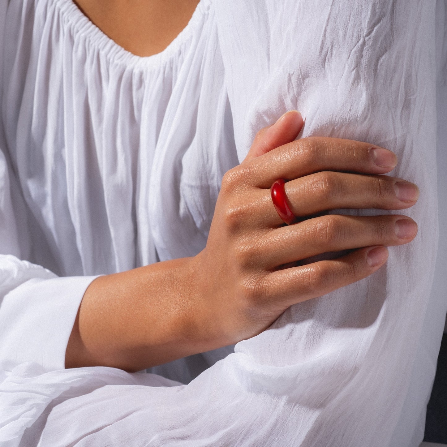 A person crosses their arms in a flowy white blouse, showcasing the Persimmon Dark Orange Agate Stone Ring on their finger. The image highlights the ring and hands with soft natural lighting.