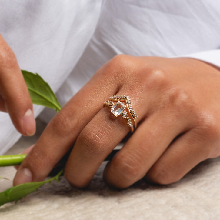 A hand wears the 14k Vintage Inspired Majesty Ring, featuring a large clear gemstone and small diamonds, paired with a matching curved band, resting on a light surface while holding a green leaf.