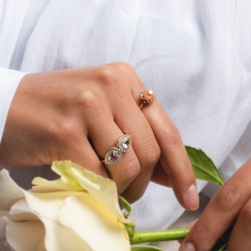 A hand in a white garment holds a pale yellow rose, wearing the 14k Pastel Pink Pearl Statement Ring—a solid gold ring featuring a round pearl encircled by small stones—alongside another with three teardrop-shaped gems.