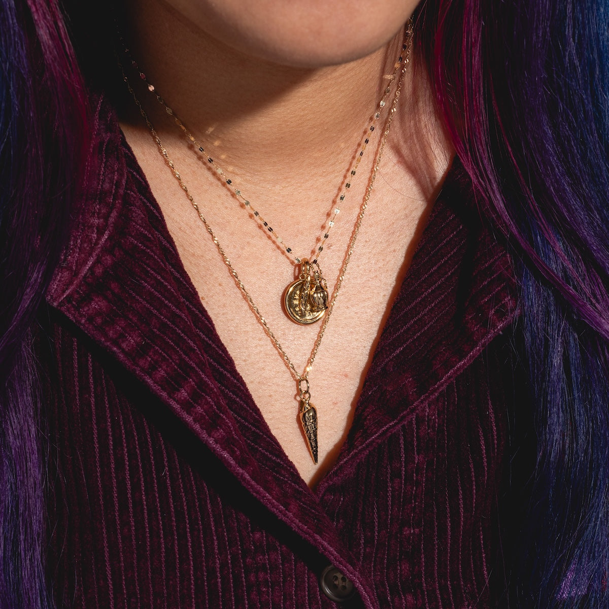 A person with long, wavy, dark hair dyed purple and blue wears Charms by I Like It Here Club—layered necklaces with 12k yellow gold plating and small charms—adding flair over a dark burgundy corduroy shirt.