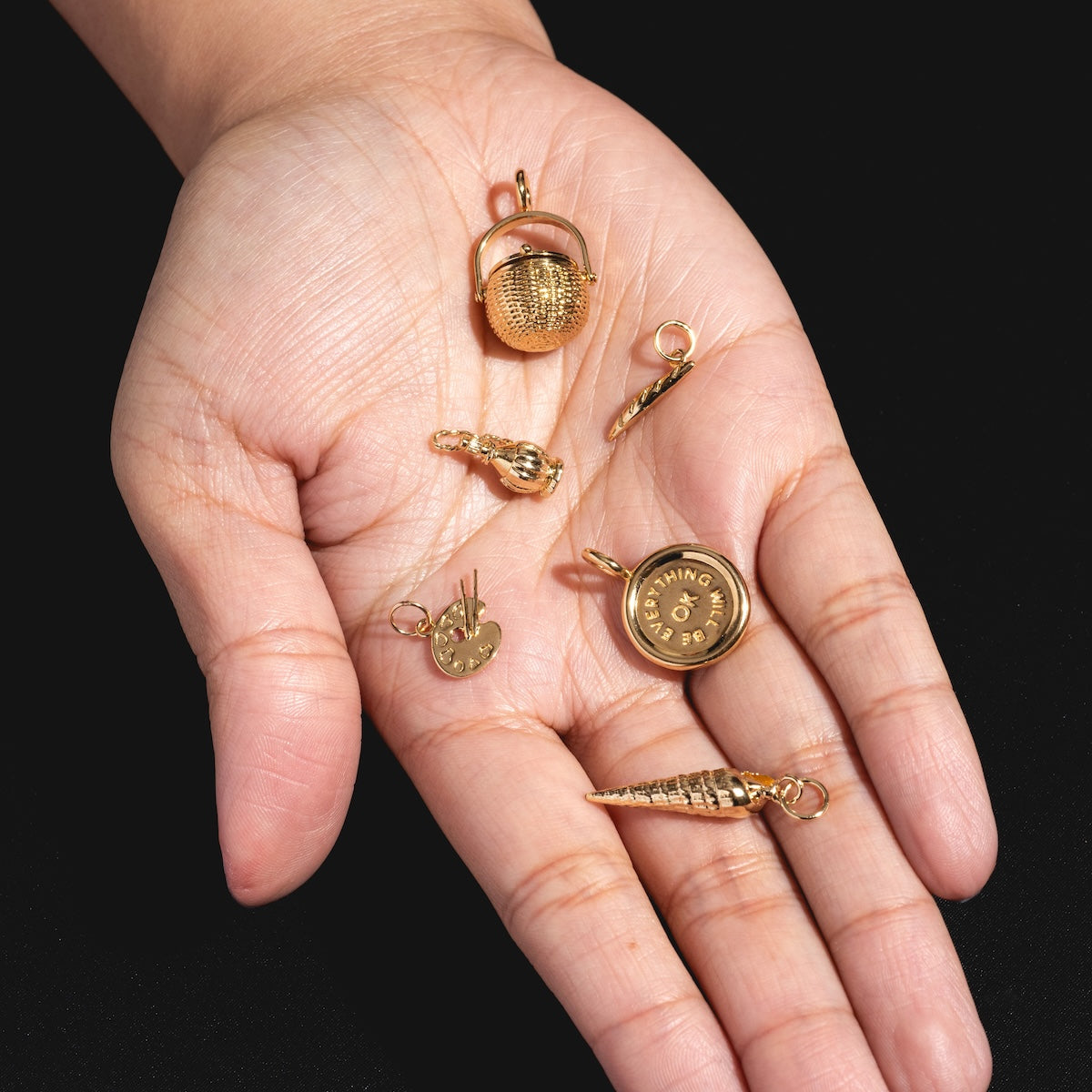 A hand displays six Charms by I Like It Here Club—basketball, number five hand sign, record disc, heart, safety pin, and spiral—all shining in 12k yellow gold plating against a black background.