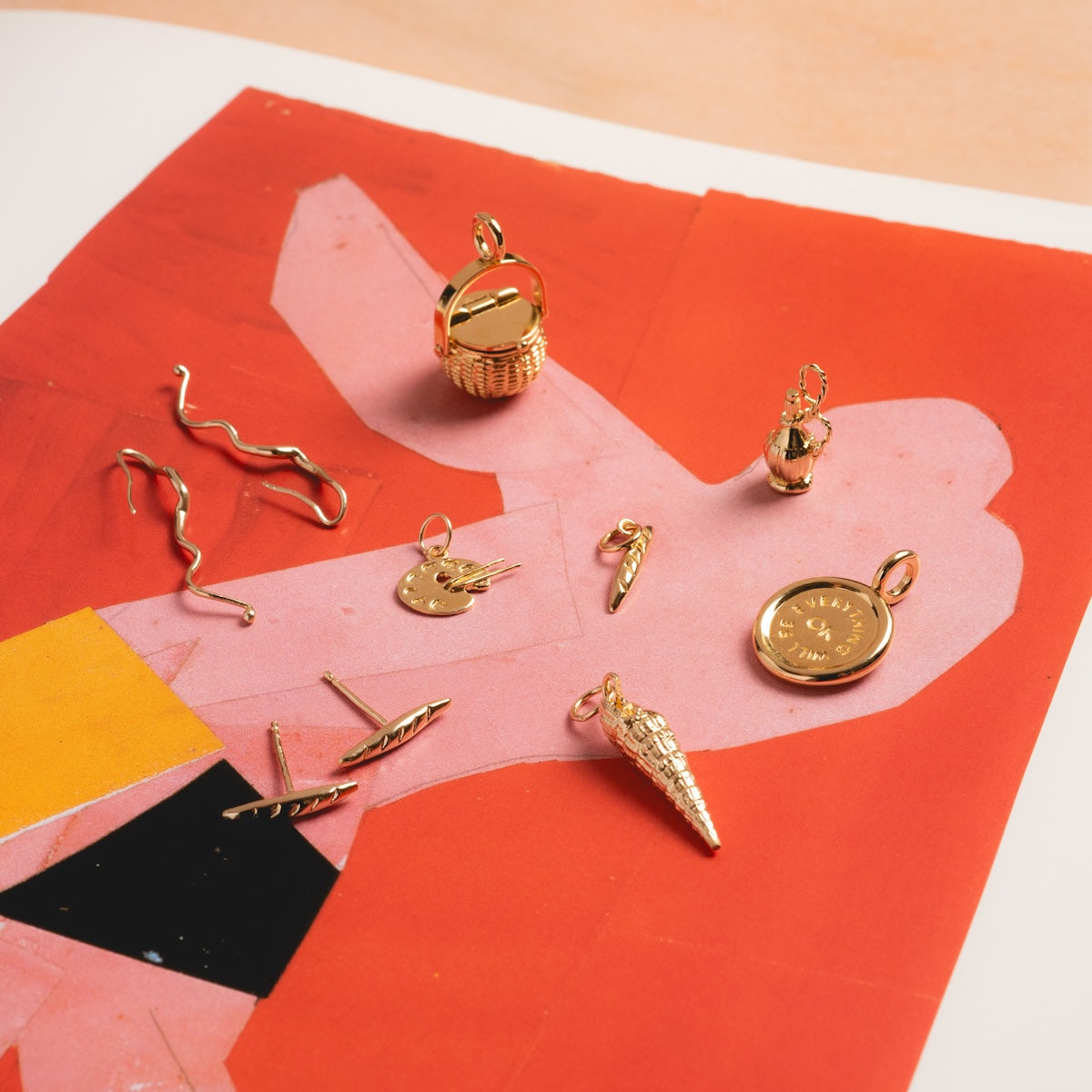 Line Study Earrings, a set of small gold-plated earrings and charms in whimsical shapes like a basket, ice cream cone, hand, and feather, are displayed on a vibrant red and pink abstract background.