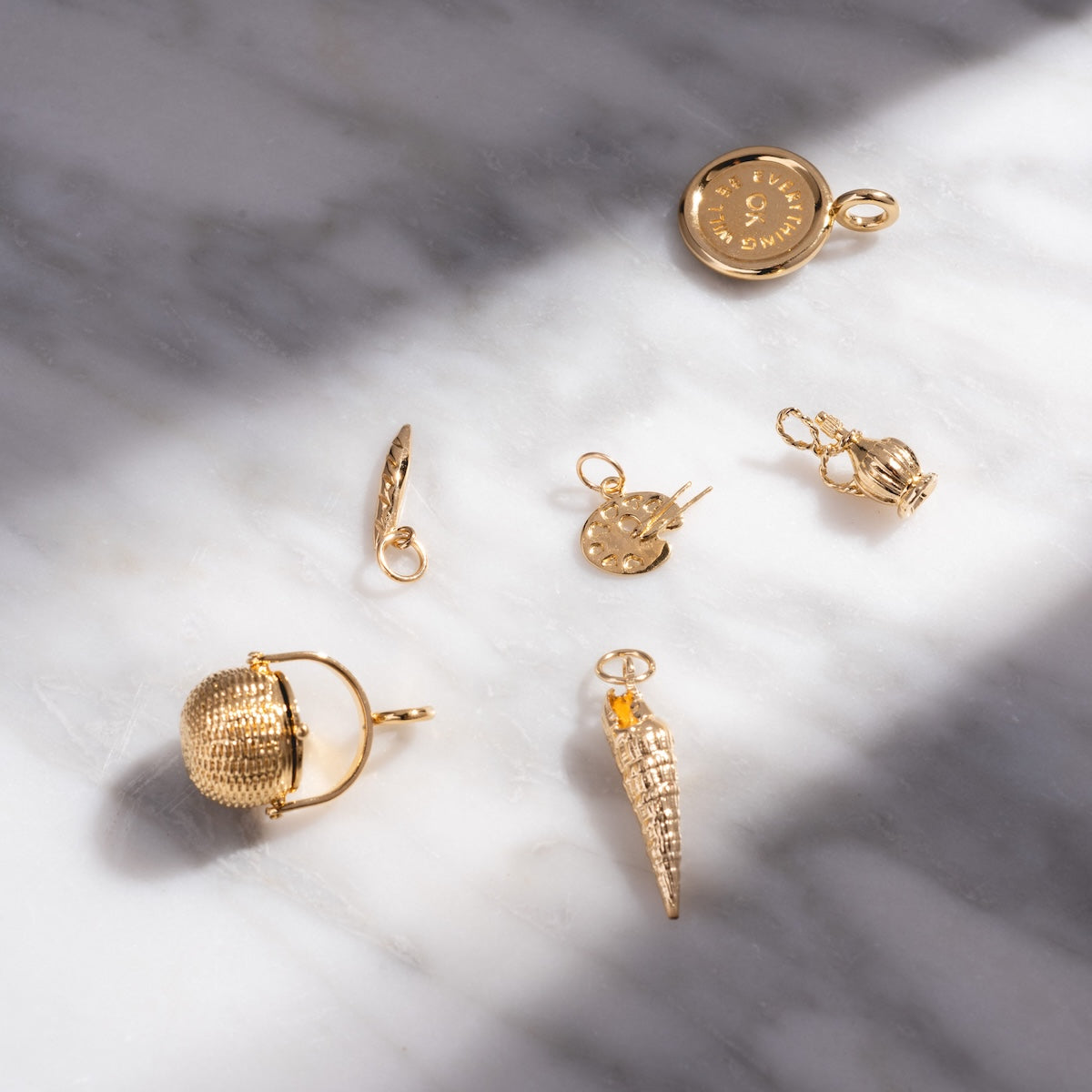 Six small Charms by I Like It Here Club, plated in 12k yellow gold—a compass, spiral shell, disc, oval, crown, and textured sphere—are artfully arranged on marble with light and shadow for a personalized touch.