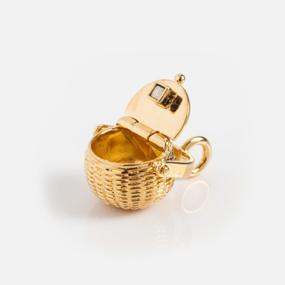 The Charms by I Like It Here Club features a small basket-shaped locket with 12k yellow gold plating, a hinged lid revealing a smooth interior, woven-texture exterior, and a loop for attaching to chains or bracelets.