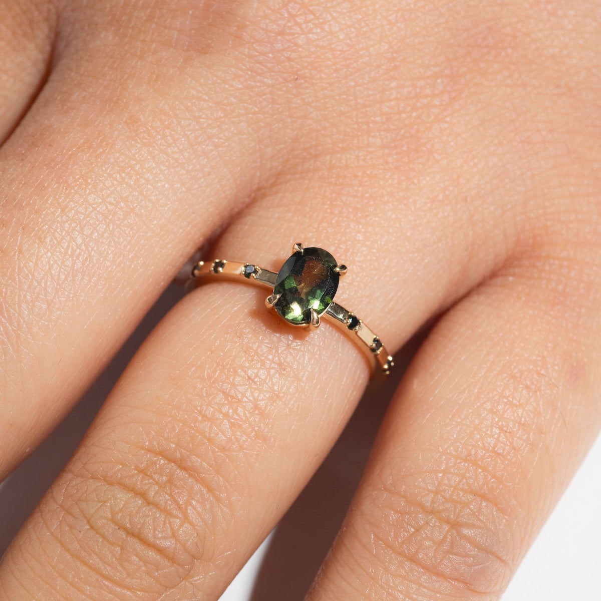 A close-up of a hand wearing the Iona Ring Black Diamond Ring, crafted in 14k yellow gold with a green oval gemstone and small black diamonds set along the band.