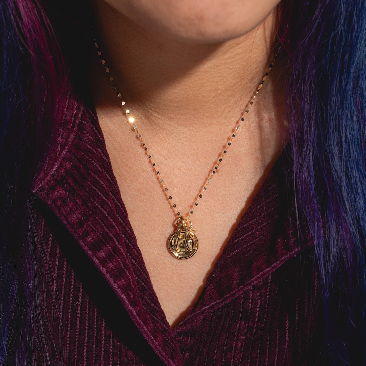 A person with long, dark purple hair wears a ribbed burgundy top and Charms by I Like It Here Club—a face-design pendant necklace with 12k yellow gold plating—adding a personalized touch to their elegant look.