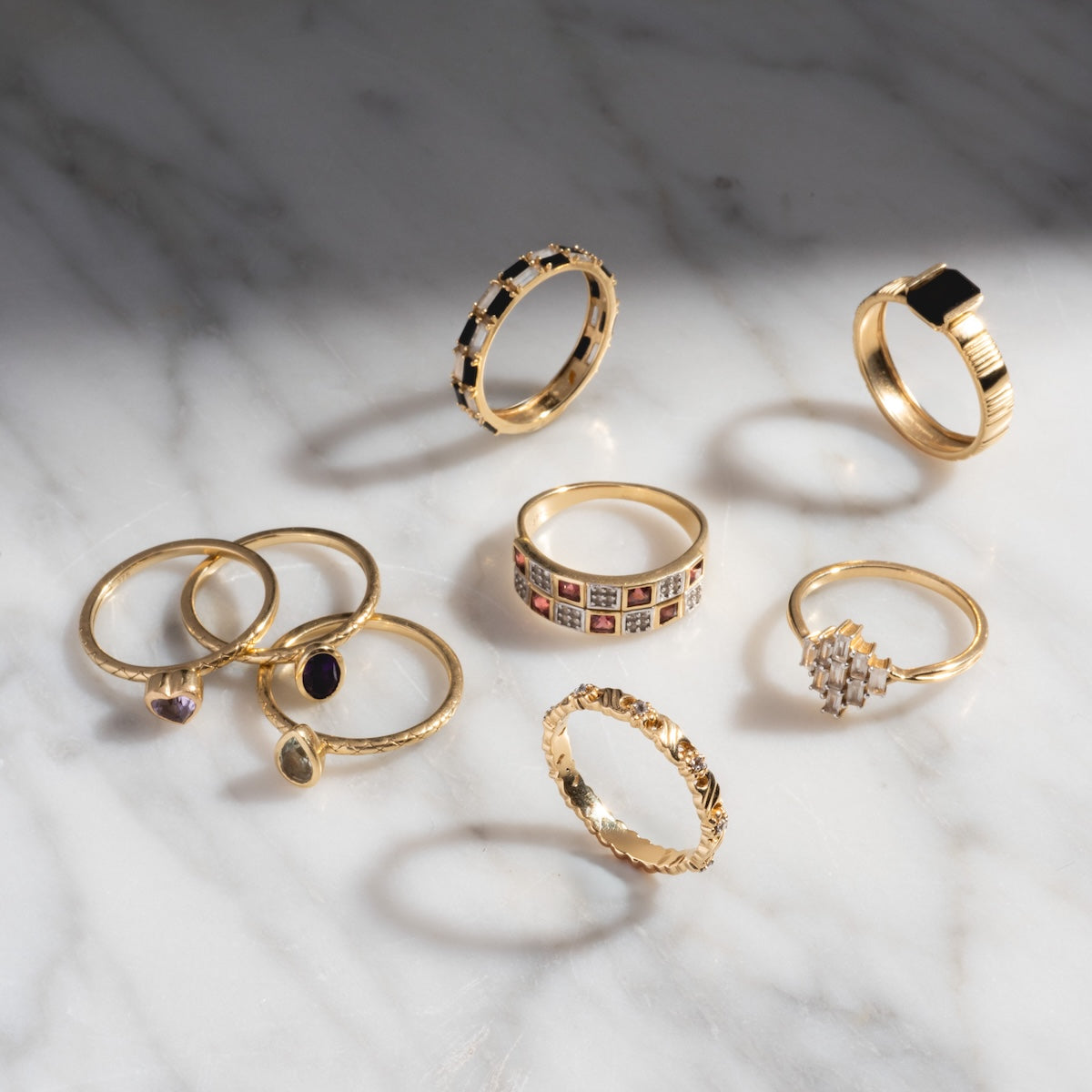 Seven Family Gold Solid Gold Garnet and Topaz Checkerboard Bands are displayed on a white marble surface, their garnet and topaz accents casting elegant shadows in natural light.
