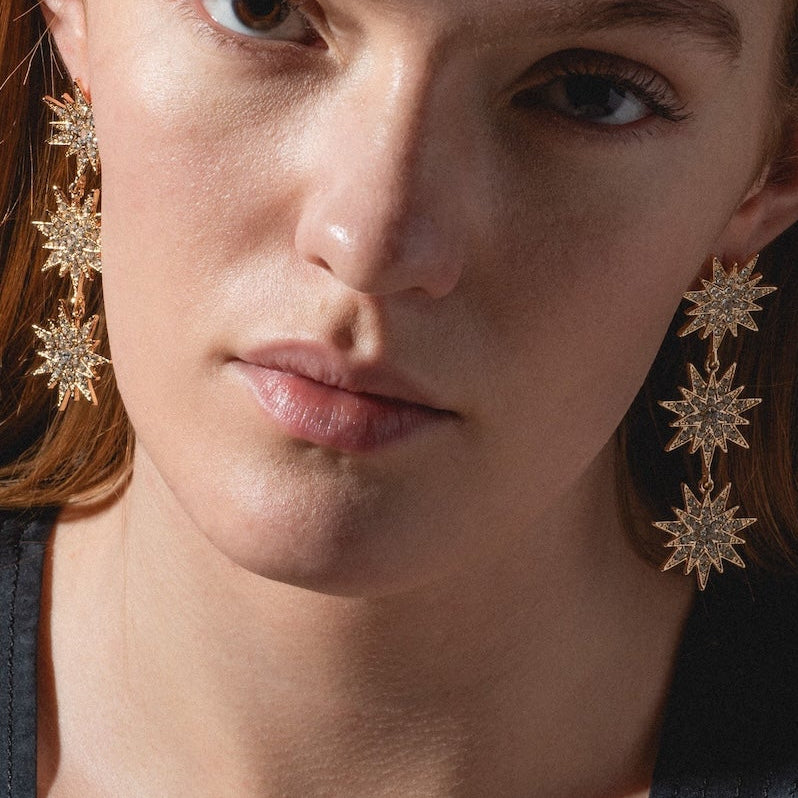 A woman with long red hair and a black top wears Celestial Drop Earrings—large, gold, star-shaped dangles—while looking directly at the camera with a neutral expression.