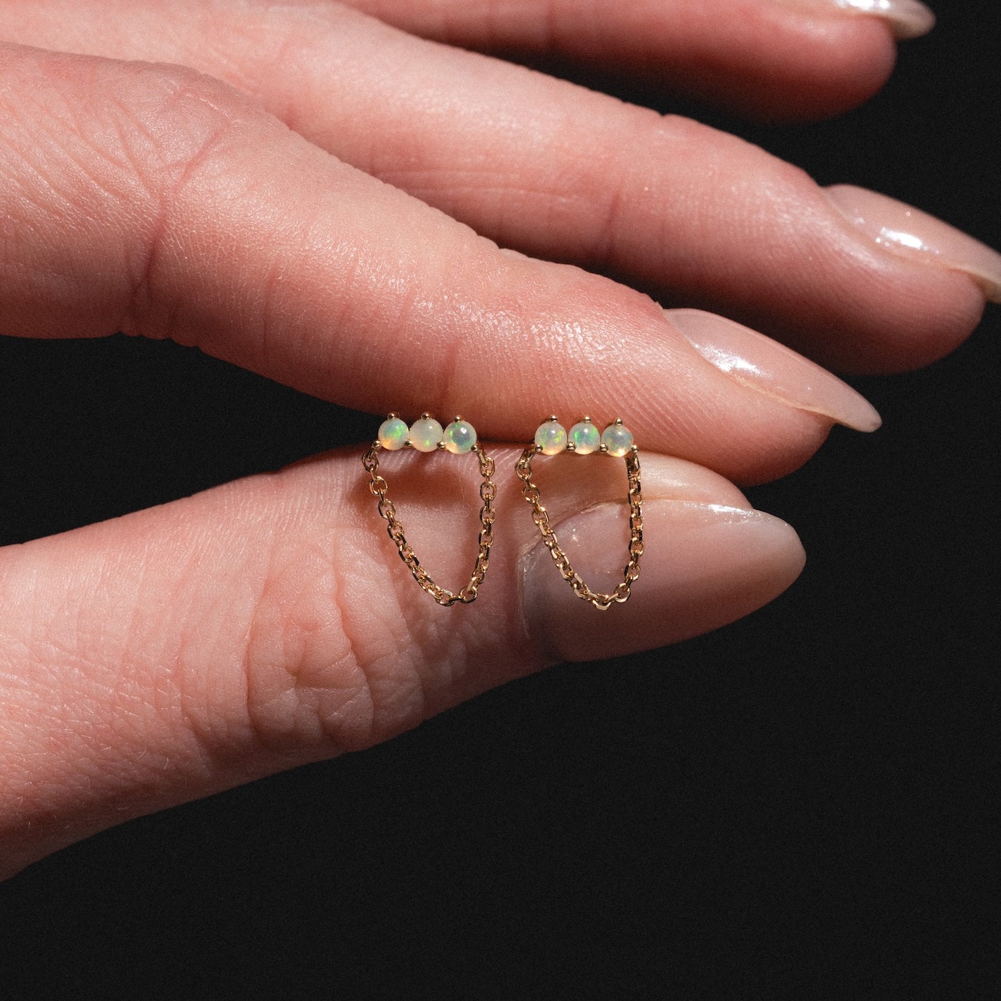 A manicured hand holds the Solid Gold Opal Trio Chain Dangle Studs—each earring features three Ethiopian opal beads linked by a delicate gold chain—shown against a black background.