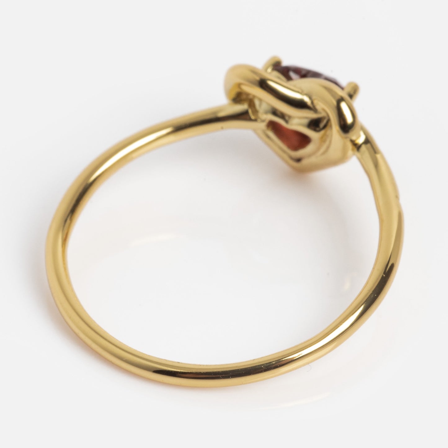 The Lover's Knot Heart Ring features a simple gold band and a single oval red gemstone set in an artistic, intertwined prong setting, photographed against a white background.