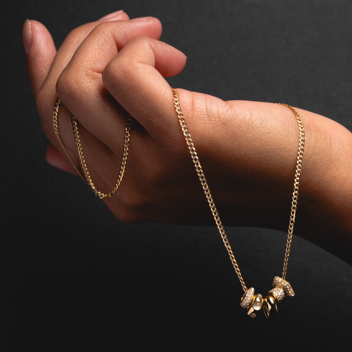 A hand holds a gold chain necklace featuring three Solid Gold Spacer Charms against a black background.