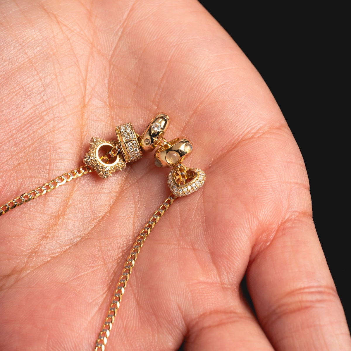 A close-up of a persons hand holding a delicate gold chain necklace featuring two gem-encrusted heart pendants and a Solid Gold Spacer Charm, all set against a black background.