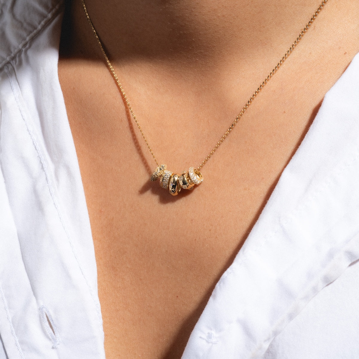 A close-up shows a person in a white collared shirt wearing Family Golds Solid Gold Spacer Charm necklace, featuring 9k yellow gold with sparkling pendants and gemstone charm spacers resting against their skin.