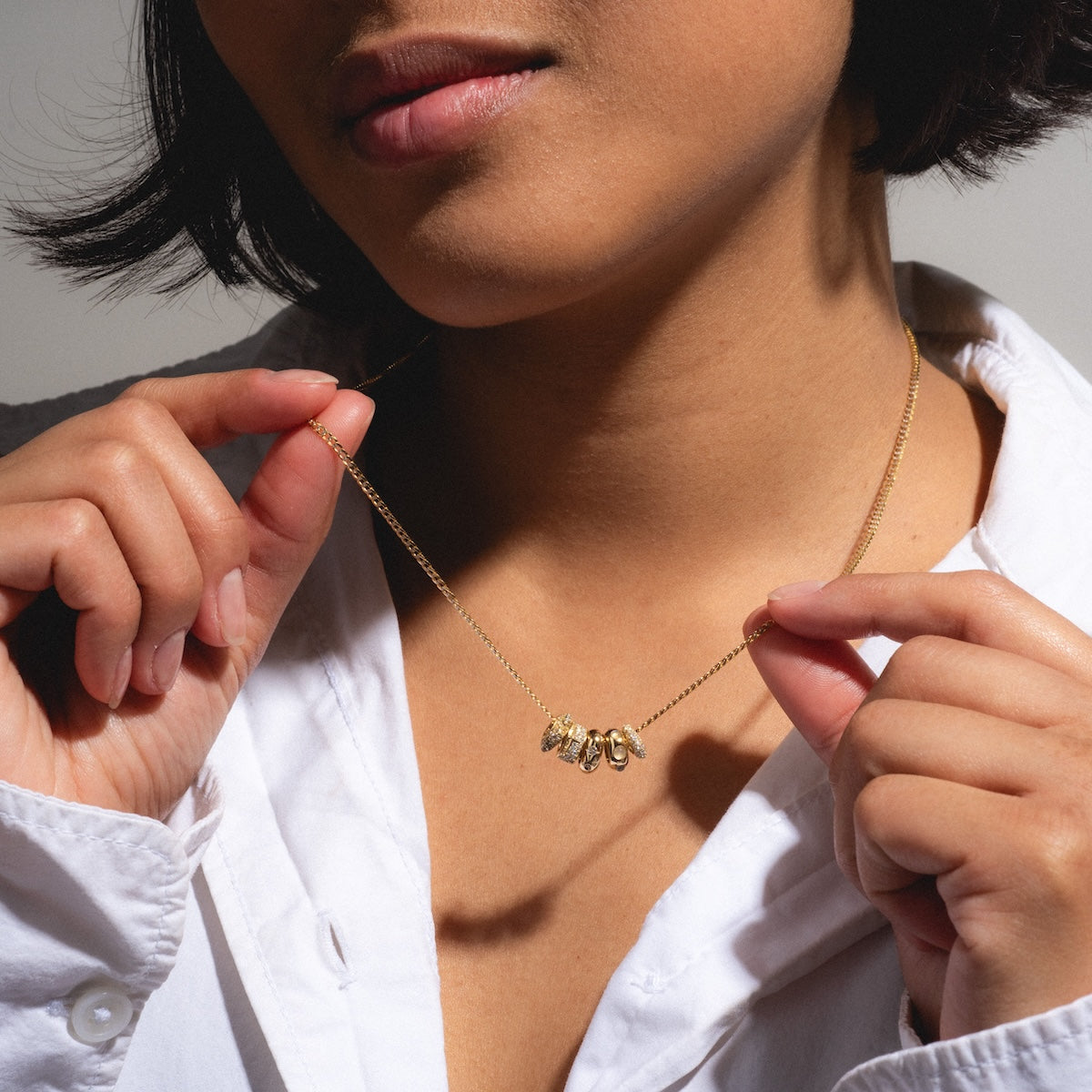 A person in a white shirt holds up a Solid Gold Spacer Charm, featuring multiple pendants in yellow gold, showcasing it near their neck with their face partially visible and the focus on the jewelry.