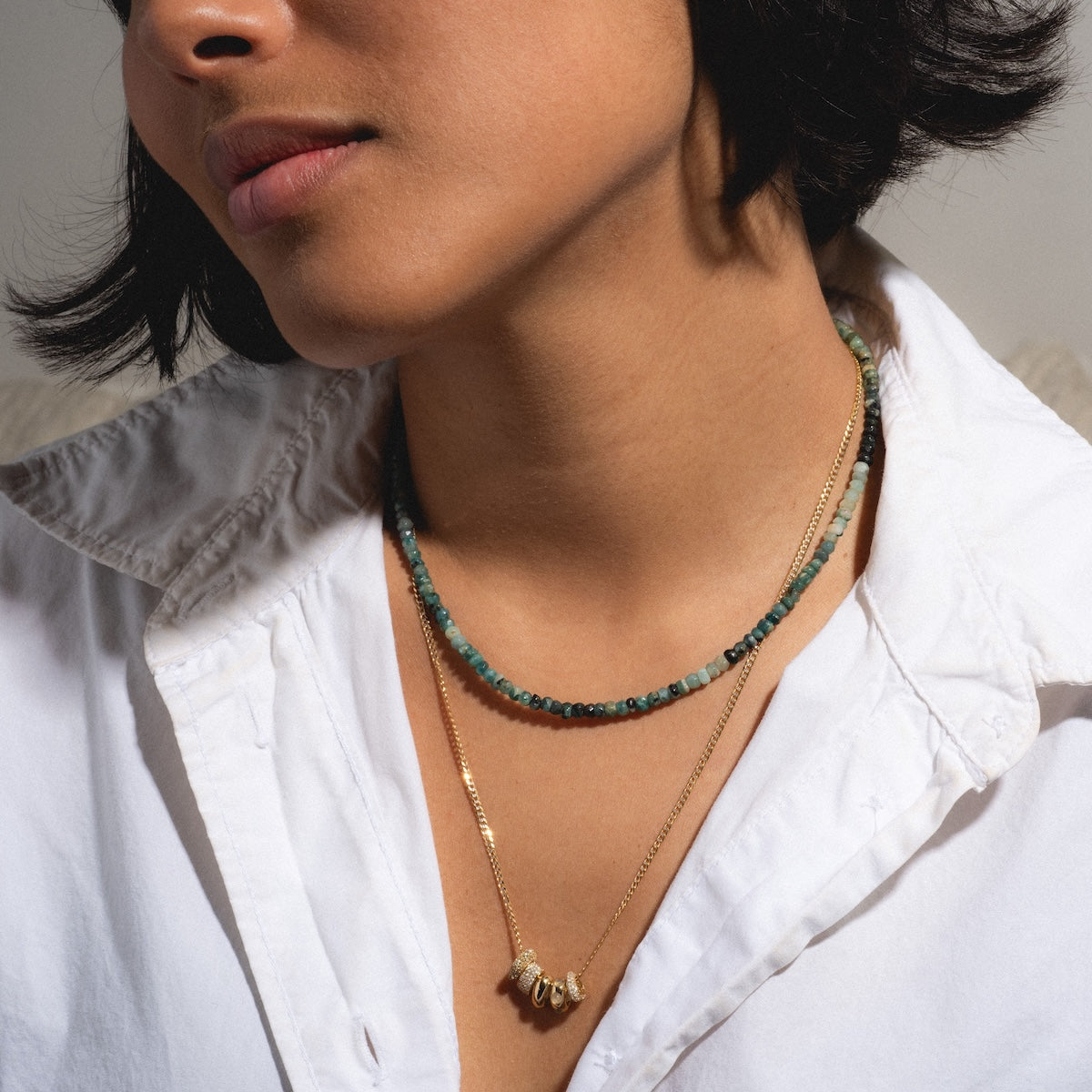 A person in a white collared shirt wears two layered necklaces: one beaded with green stones and Solid Gold Spacer Charms, the other a gold chain with curved yellow gold charms. The persons face is partially visible.