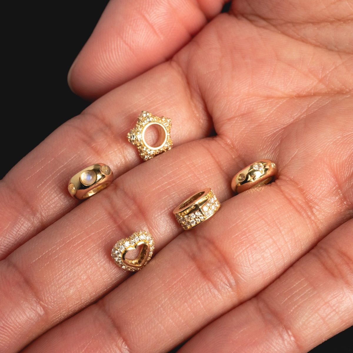 A hand displays five Solid Gold Spacer Charms nose rings, each featuring a unique design such as hearts, stars, and clear gemstones, all set against a black background.
