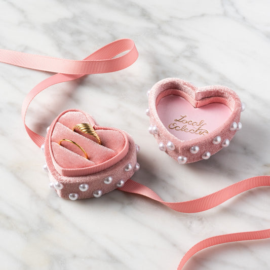 A Heart Shaped Velvet Pearl Ring Box with pearls sits open on marble, showing two gold rings inside. A pink ribbon is wrapped around the box, and Lovel, Colette is written in gold inside the lid.