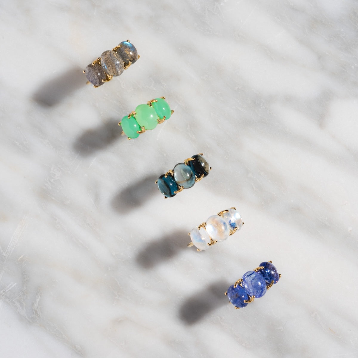 Five Sweetest Gumdrop Rings in yellow gold, featuring natural gemstones in gray, green, dark teal, white, and blue, are arranged diagonally on a white marble surface.